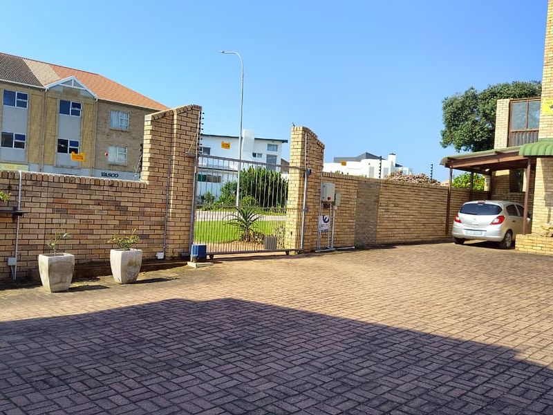 Front exterior view of the townhouse featuring a paved driveway, secure brick boundary wall with electric fencing, and a covered carport area.