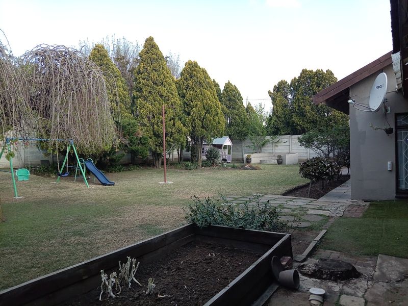 A spacious rear garden featuring a lawn, mature trees, a stone pathway, and a raised timber planter box.