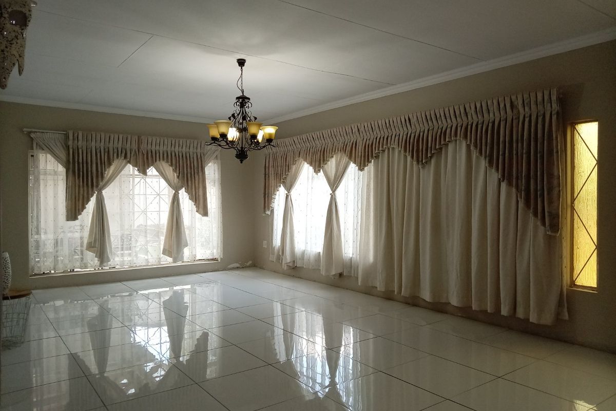 Spacious living area featuring polished tiled flooring, decorative chandelier lighting, and large windows with integrated security bars and traditional curtain pelmets.