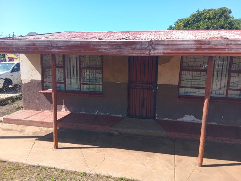 Front elevation of a single-story house featuring a corrugated iron roof, security gate, and burglar bars, showing signs of significant exterior wear and weathering.