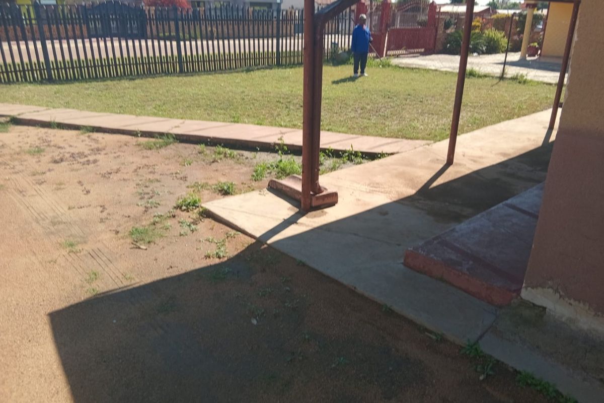 Rear exterior view showing a concrete patio area, metal support posts, and a fenced garden space with visible wear on the building exterior.