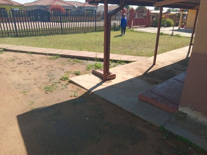 Rear exterior view showing a concrete patio area, metal support posts, and a fenced garden space with visible wear on the building exterior.