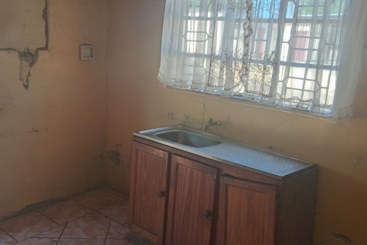 A basic kitchen area featuring a single stainless steel sink unit with wooden cabinetry, tiled flooring, and security bars on the window.