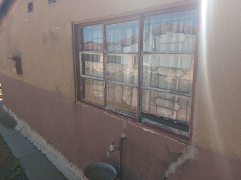 Exterior view of a residential wall featuring a sliding window with security bars and an external water tap, showing signs of significant wear.