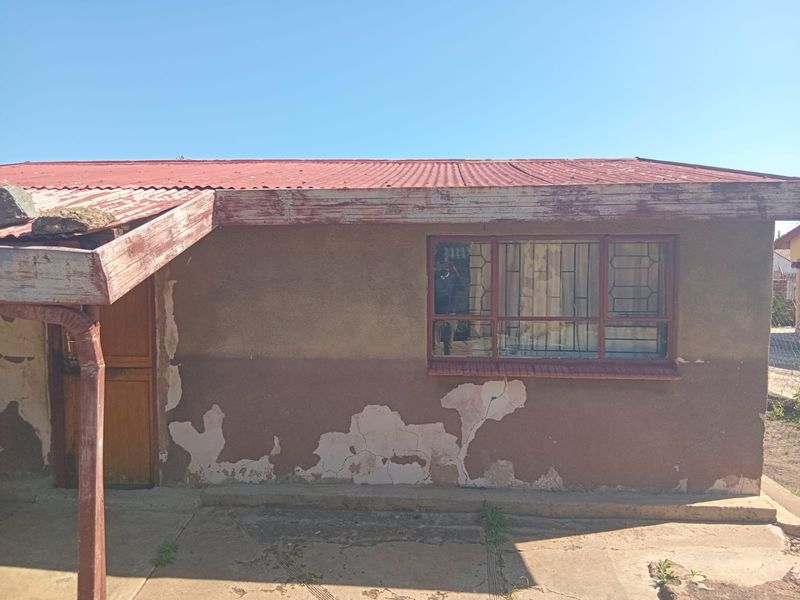 Front elevation of a house featuring a corrugated metal roof, security bars on windows, and significant exterior wall finish deterioration requiring maintenance.