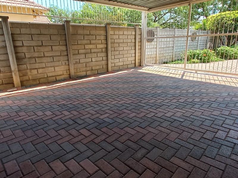 A covered carport area featuring herringbone pattern paving, enclosed by a concrete block boundary wall and a secure metal gate.
