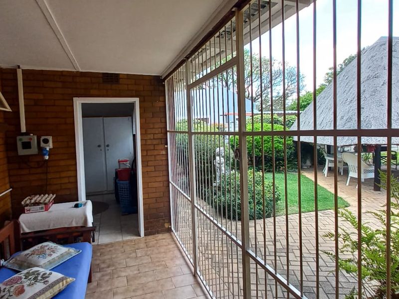 A covered patio area featuring exposed brick walls, tiled flooring, and security bars, overlooking a garden with a thatched lapa.