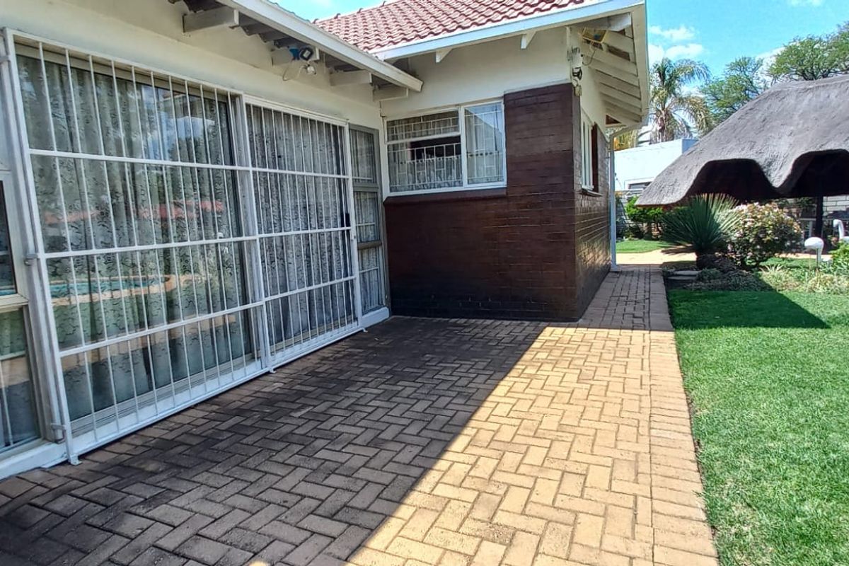 Rear exterior view featuring a paved patio area, sliding glass doors with security bars, and a thatched lapa in the garden.