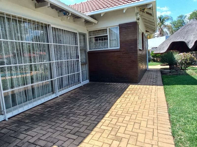Rear exterior view featuring a paved patio area, sliding glass doors with security bars, and a thatched lapa in the garden.