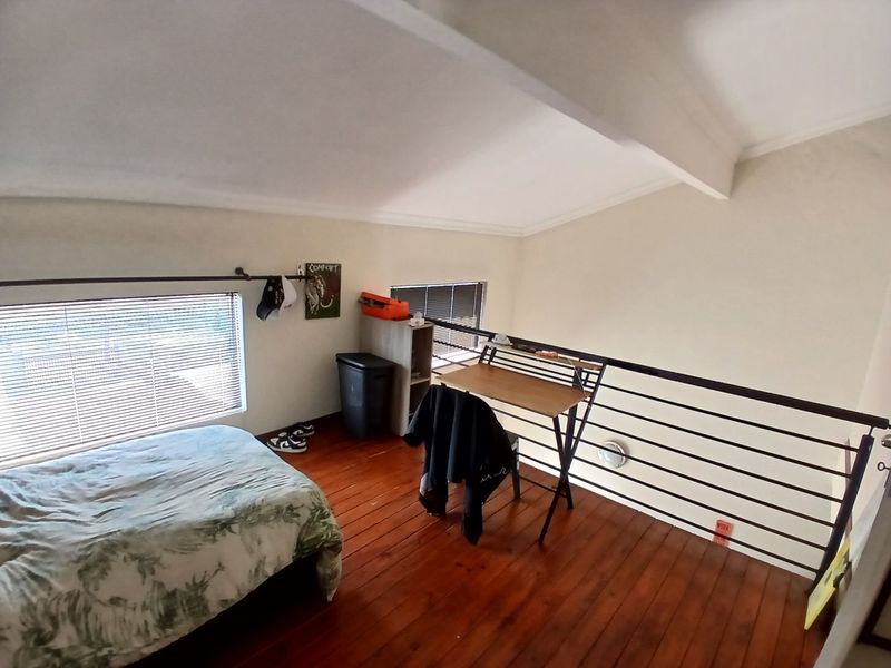 A loft-style bedroom featuring timber flooring, a metal safety balustrade, and a sloped ceiling with natural light from venetian blinds.
