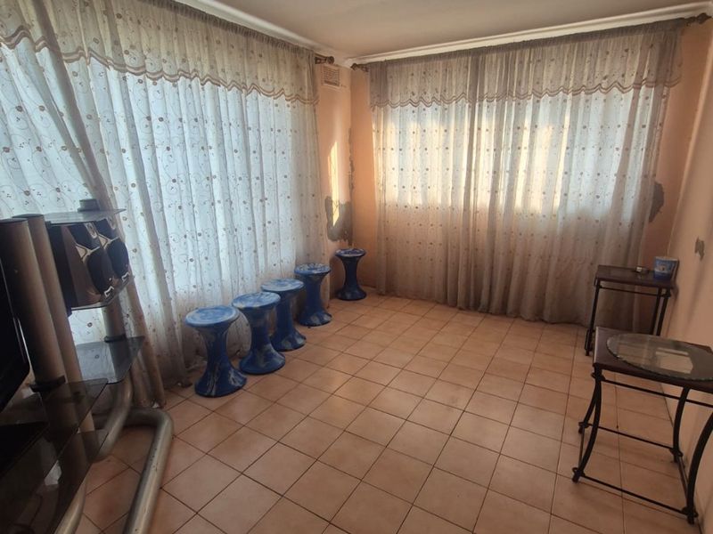 A living room featuring tiled flooring and large windows with curtains, showing visible signs of wall surface wear and peeling paint.