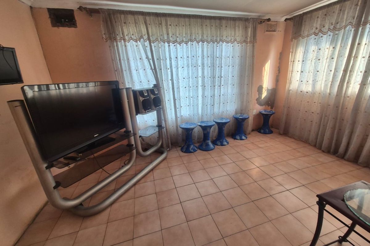 A living room featuring tiled flooring and large windows with curtain rails, currently showing signs of wall plaster damage and paint deterioration.