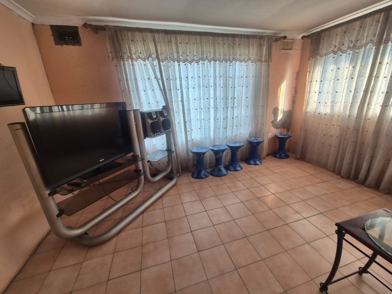 A living room featuring tiled flooring and large windows with curtain rails, currently showing signs of wall plaster damage and paint deterioration.