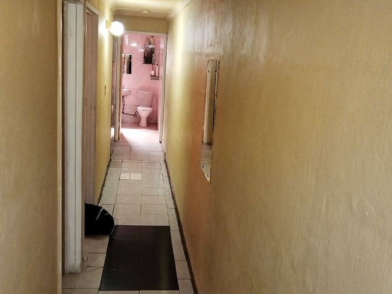A narrow residential hallway with tiled flooring leading towards a bathroom, featuring plain walls and basic architectural finishes.