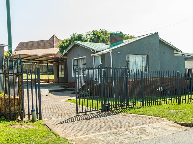 Front view of a single-story house featuring a paved driveway, carport, metal security fencing, and a brick chimney.