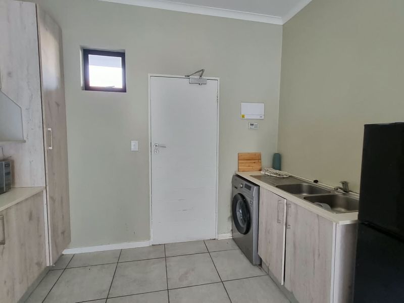 A functional kitchen featuring light wood-look cabinetry, built-in oven and hob, stainless steel sink, and tiled flooring with direct access to an exterior door.