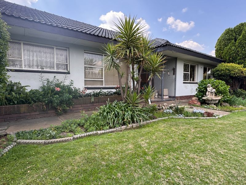 Front elevation of a single-story house featuring a tiled roof, security gates, and established garden beds with a concrete pathway.