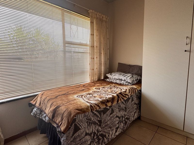 A functional bedroom featuring tiled flooring, a built-in wardrobe, and large windows fitted with venetian blinds.