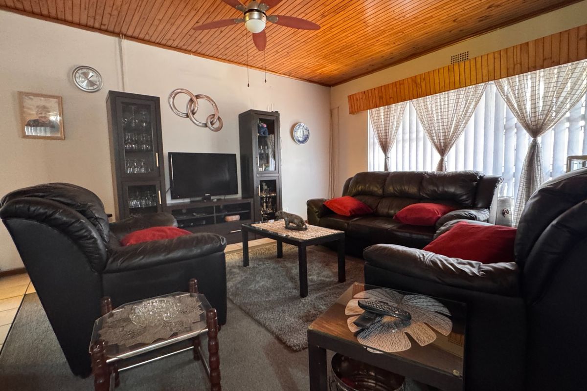 A comfortable living room featuring a warm timber tongue and groove ceiling, ceiling fan, and tiled flooring with large windows and vertical blinds.