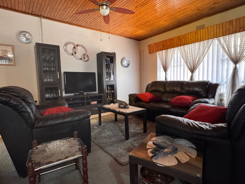 A comfortable living room featuring a warm timber tongue and groove ceiling, ceiling fan, and tiled flooring with large windows and vertical blinds.