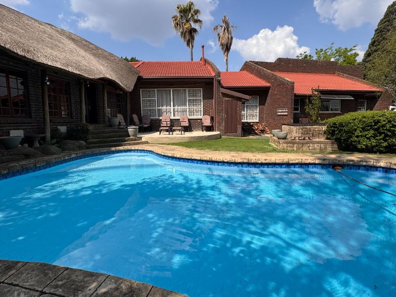 A spacious outdoor pool area featuring a swimming pool, paved patio, and a mix of traditional thatched and tiled roof architecture.