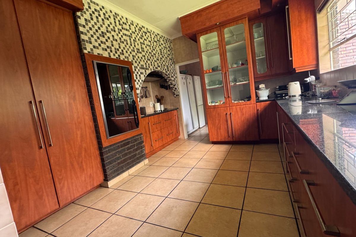 A spacious kitchen featuring warm-toned wooden cabinetry, granite countertops, tiled flooring, and a unique brick and mosaic tile feature wall.