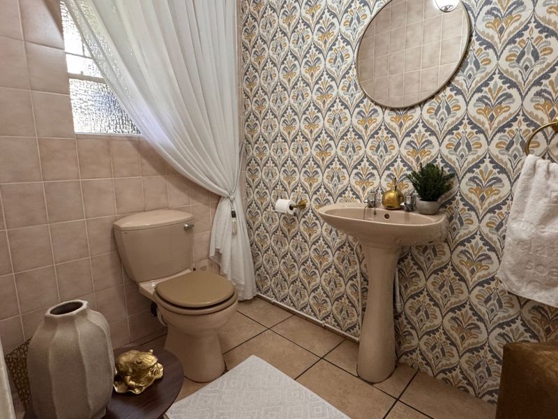 A compact bathroom featuring a pedestal basin, toilet, tiled walls, and patterned wallpaper, offering a functional space with a dated aesthetic.