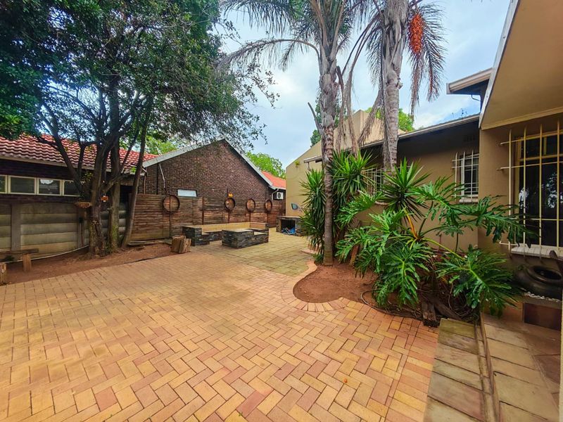 A rear garden area featuring a paved patio, built-in braai, and established landscaping with mature trees and shrubs.