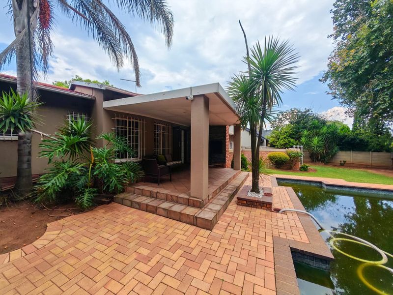 A covered patio area featuring brick paving and a built-in fireplace, overlooking a swimming pool in the rear garden.
