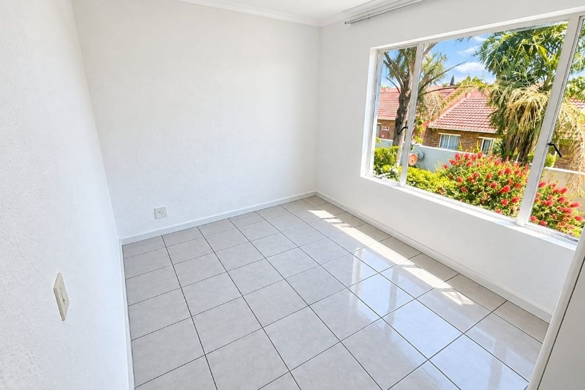 A bright, empty bedroom featuring neutral tiled flooring and a large window providing natural light and a pleasant garden view.