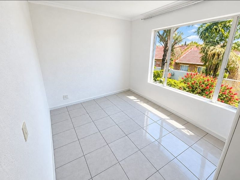 A bright, empty bedroom featuring neutral tiled flooring and a large window providing natural light and a pleasant garden view.