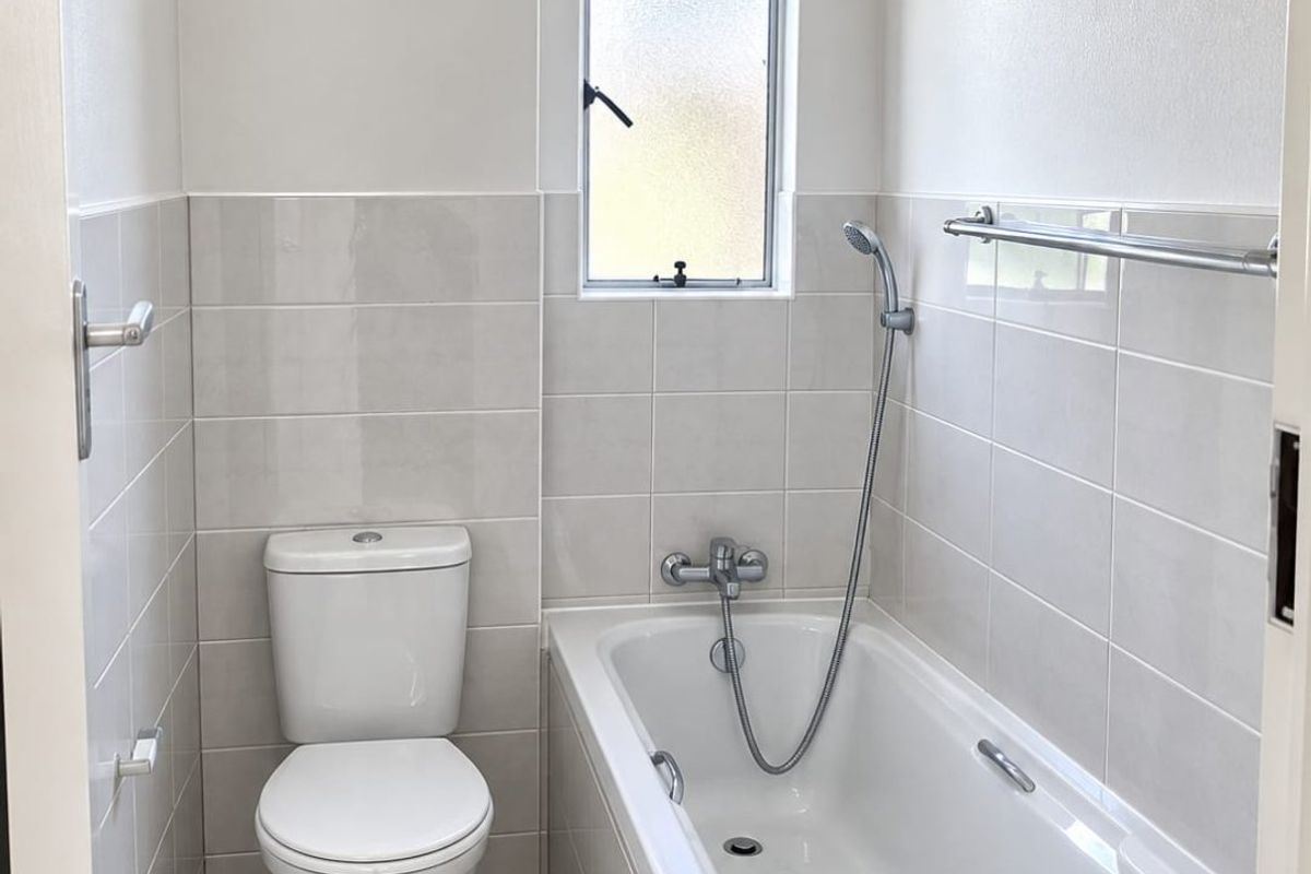 A clean, modern bathroom featuring a white bathtub with shower mixer, a standard toilet, and neutral wall and floor tiling.