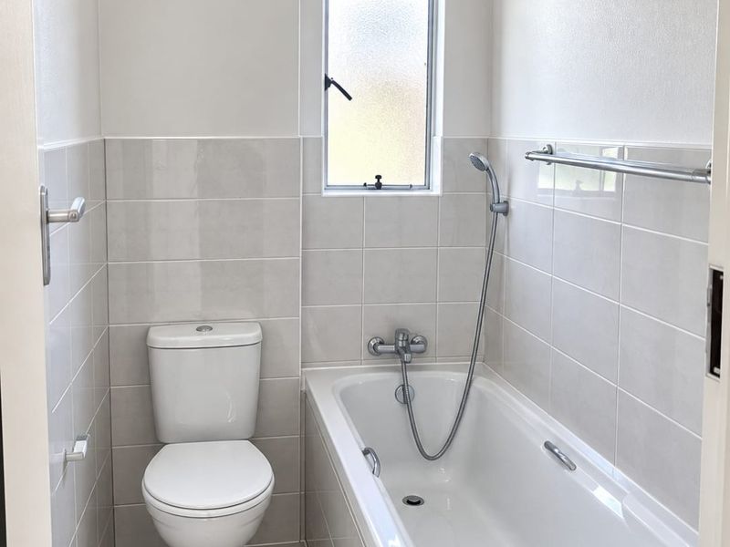 A clean, modern bathroom featuring a white bathtub with shower mixer, a standard toilet, and neutral wall and floor tiling.
