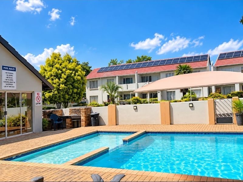 Communal swimming pool area featuring brick paving, a built-in braai, and a shade sail, set within a well-maintained apartment complex with solar panels.