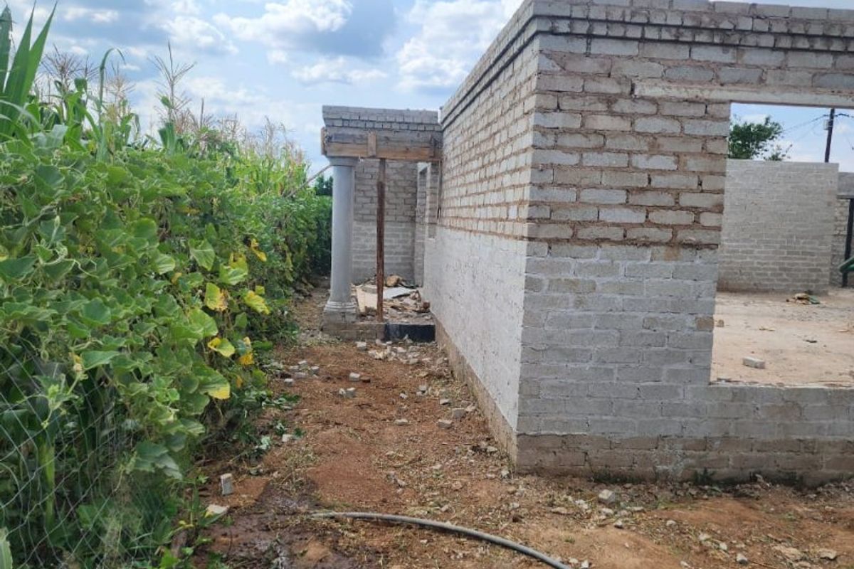 An exterior view of a house under construction, featuring exposed brick walls and a concrete support column in a rural setting.