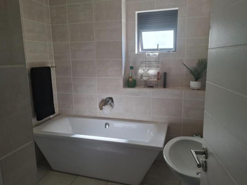 A modern bathroom featuring a white bathtub with wall-mounted fixtures, neutral wall tiling, and a built-in recessed shelf under a window.