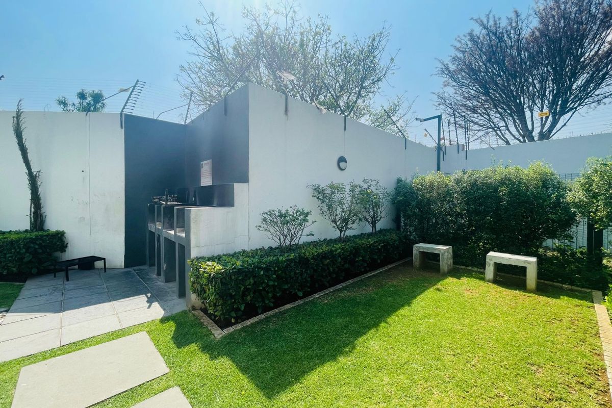 A private rear garden area featuring a built-in braai, paved pathways, manicured lawn, and concrete seating, secured by perimeter electric fencing.