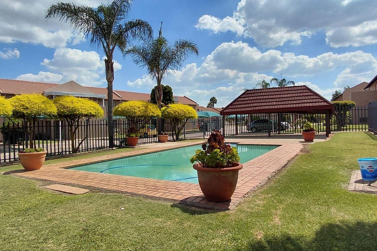 Communal outdoor pool area featuring a swimming pool with brick coping, surrounding lawn, and a covered gazebo structure within a fenced complex.
