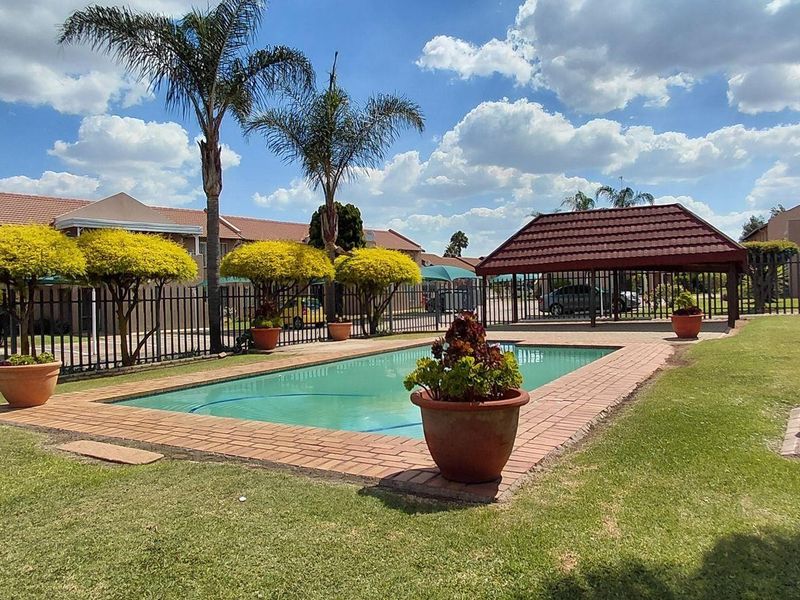 Communal outdoor pool area featuring a swimming pool with brick coping, surrounding lawn, and a covered gazebo structure within a fenced complex.