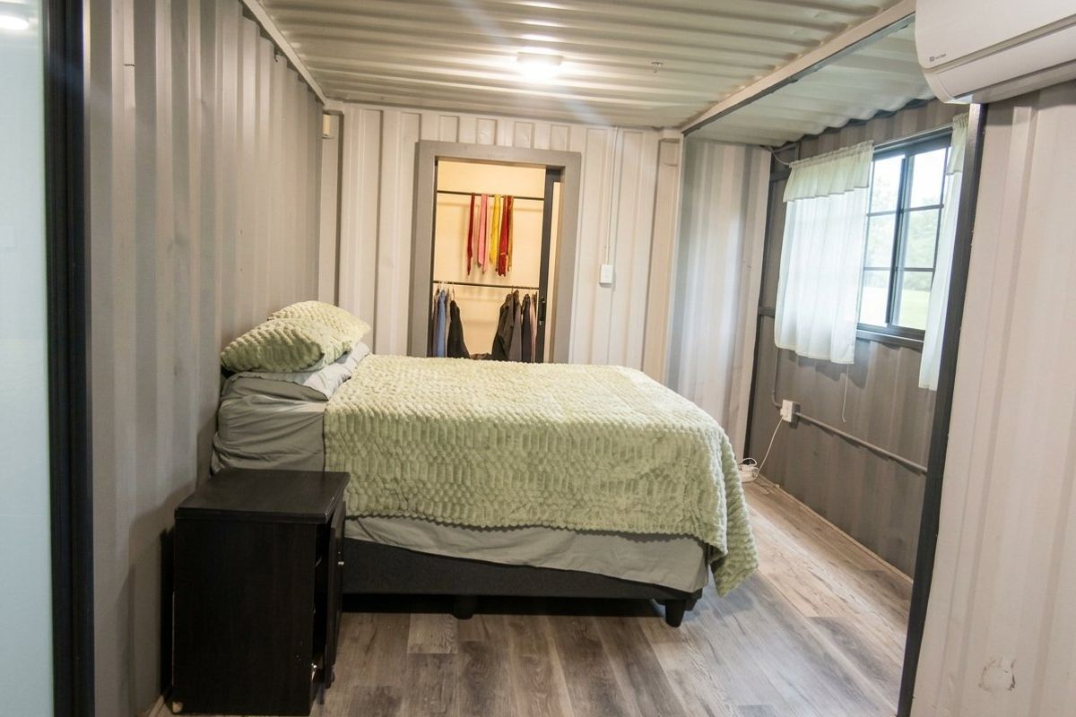 A unique bedroom constructed from a shipping container, featuring corrugated metal walls, laminate flooring, and an integrated split-system air conditioner.