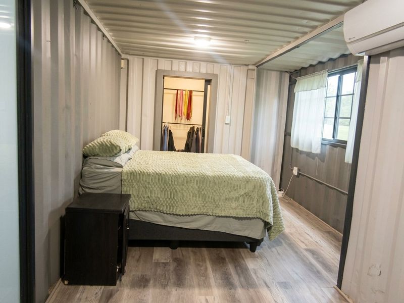 A unique bedroom constructed from a shipping container, featuring corrugated metal walls, laminate flooring, and an integrated split-system air conditioner.