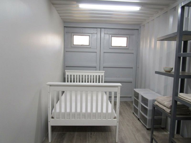 A bedroom converted from a shipping container, featuring corrugated metal walls, laminate flooring, and integrated container doors with small windows.