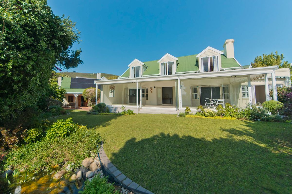 Rear view of a well-maintained house featuring a green metal roof, dormer windows, and a spacious enclosed patio overlooking a landscaped garden and pond.