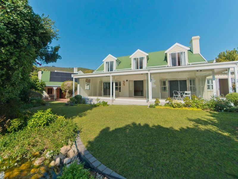 Rear view of a well-maintained house featuring a green metal roof, dormer windows, and a spacious enclosed patio overlooking a landscaped garden and pond.