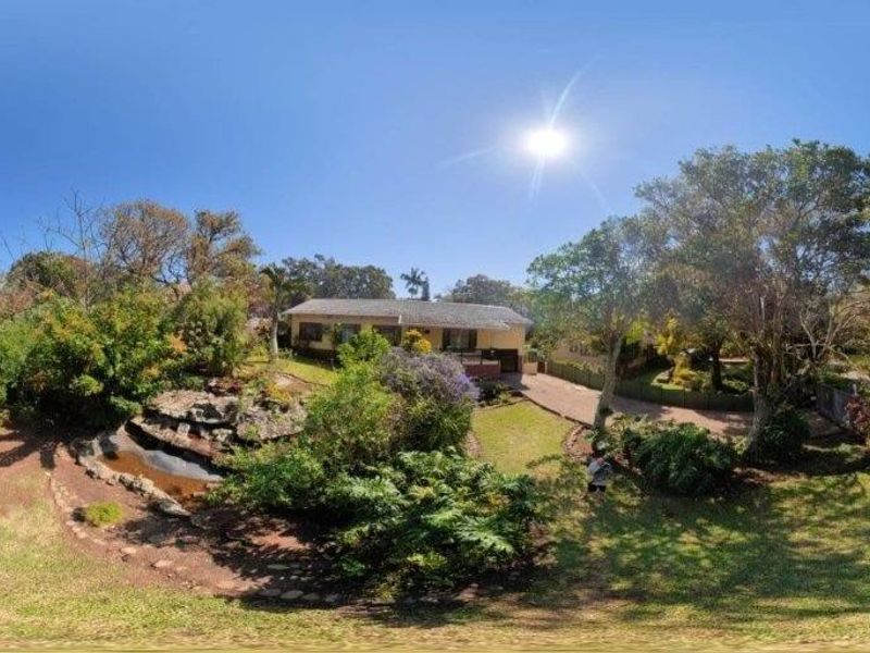 A panoramic view of the front exterior of the house, featuring a paved driveway, established garden, and a decorative rock water feature.