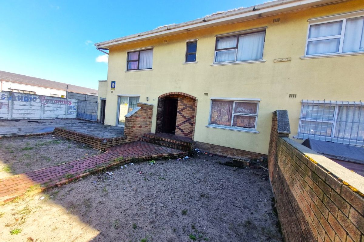 Rear view of a two-story residential building featuring a paved patio, brick boundary walls, and aluminum-framed windows requiring exterior maintenance.