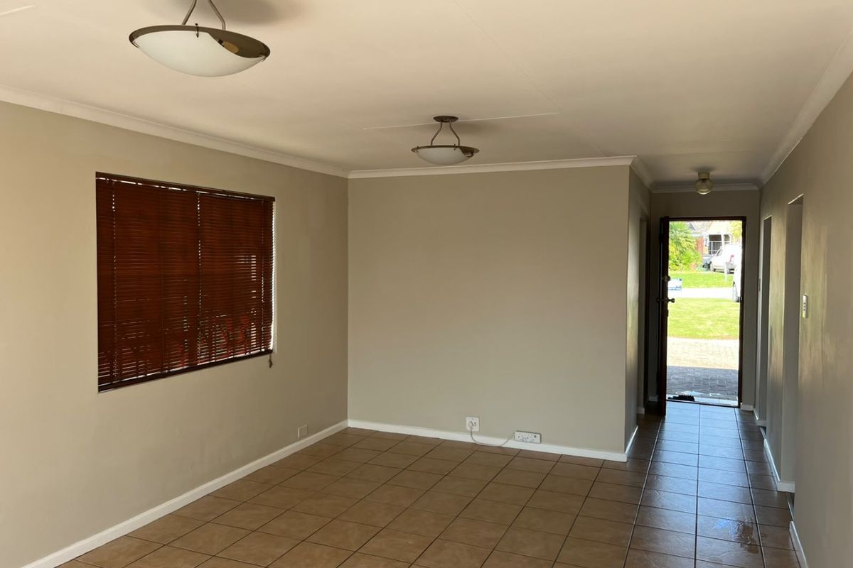 An empty, open-plan living area featuring neutral wall tones, ceramic tiled flooring, and natural light through a window with fitted venetian blinds.