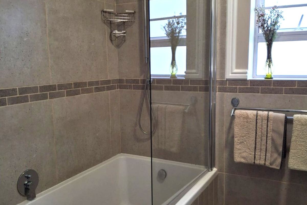 A well-maintained bathroom featuring a built-in bathtub with a glass shower screen, neutral wall tiling, and natural light from the windows.