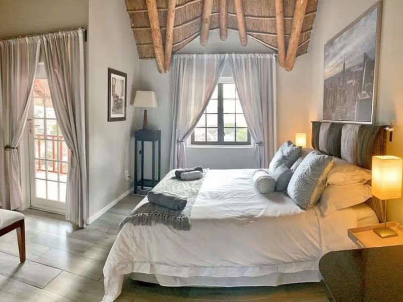 A well-maintained bedroom featuring a high vaulted ceiling with exposed timber rafters, thatched roofing, tiled floors, and direct access to a balcony via french doors.