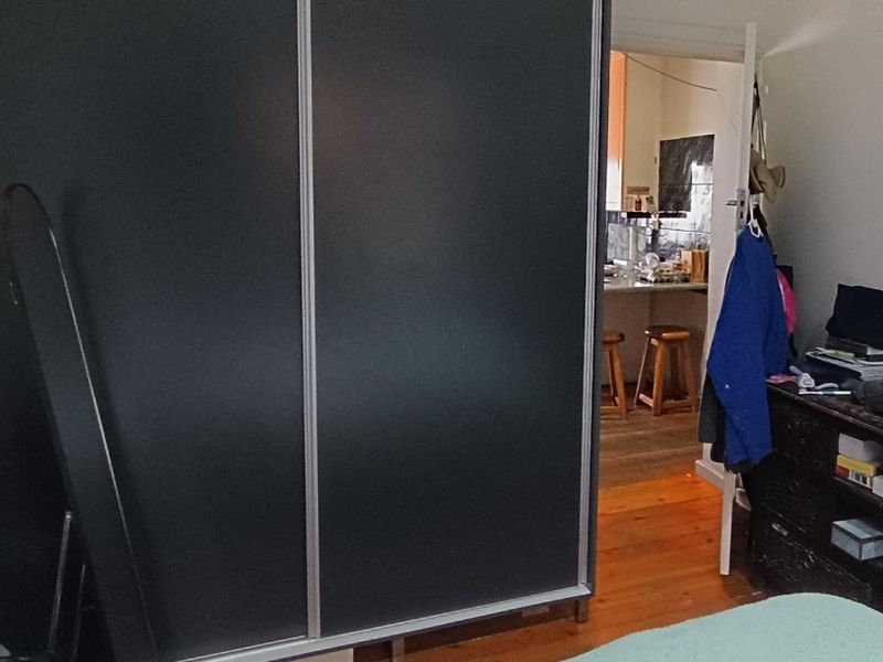 Bedroom featuring a large dark built-in wardrobe and timber flooring, with a view through the doorway into the adjacent kitchen area.